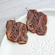 Load image into Gallery viewer, A pair of handmade copper earrings with intricate hand-carved paisley and floral patterns, resting on a white ceramic dish over a light gray wood background.