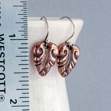Load image into Gallery viewer, A pair of copper leaf shaped dangle earrings with oxidized dark detailing, displayed on a white ceramic stand next to a metal ruler for scale against a gray background.