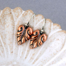 Load image into Gallery viewer, A pair of copper leaf-shaped drop earrings with oxidized dark detailing and silver fishhook wires, displayed on a textured white ceramic shell against a gray background.