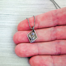 Load image into Gallery viewer, A fine silver thistle pendant with a blue tanzanite centerpiece, attached to a silver chain, held in a hand for scale