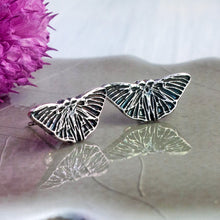 Load image into Gallery viewer, Silver butterfly-shaped stud earrings on a reflective surface with a pink flower in the background