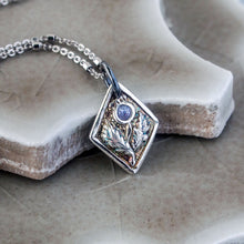 Load image into Gallery viewer, A fine silver thistle pendant with a blue tanzanite centerpiece, attached to a silver chain, displayed on a textured background.