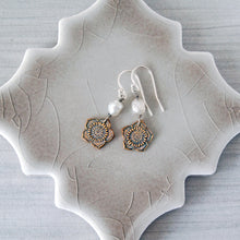 Load image into Gallery viewer, A pair of bohemian-style dangle earrings with bronze flower charms, white pearl beads, and silver fishhook wires, displayed on a textured ceramic dish over a gray wood background.