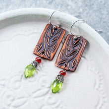 Load image into Gallery viewer, A pair of handmade dangle earrings with rectangular copper metal plates featuring leaf engravings, hanging from silver fishhook wires. Each earring has a small orange gemstone bead and a faceted green gemstone drop, resting on a white textured plate.