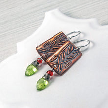 Load image into Gallery viewer, A pair of handmade dangle earrings featuring rectangular copper metal plates with engraved leaf patterns, silver fishhook wires, small red gemstone beads, and green faceted gemstones, displayed on a white surface.