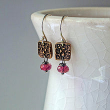 Load image into Gallery viewer, A pair of dangling earrings with textured bronze square charms, silver wire hooks, and faceted pink tourmaline beads, hanging from a white ceramic cup against a gray background.