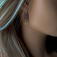 Load image into Gallery viewer, A close-up of a woman's ear wearing a single copper rectangular drop earring with engraved leaf-like detailing, a small orange gemstone bead, and a dangling green faceted gemstone, shown against her hair and skin.