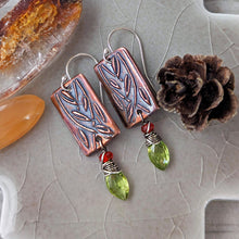 Load image into Gallery viewer, A pair of handmade rectangular drop earrings with copper metal rectangles featuring an embossed leaf pattern, silver wire hooks, small orange gemstone beads, and green faceted gemstone drops, displayed on a textured light gray stone surface with decorative props.