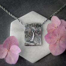 Load image into Gallery viewer, Silver pendant necklace with tree design on a white stone surface with pink flowers.