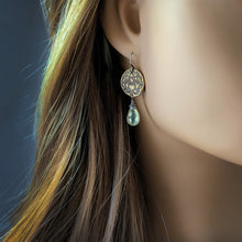 Load image into Gallery viewer, A close-up of a model's ear wearing a bronze dangle earring with an engraved circular disc and a small teardrop-shaped light green gemstone.