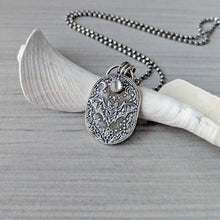 Load image into Gallery viewer, A silver necklace with an intricately designed pendant depicting a flying bat with a pale gray stone propped up against a white fossilized shell on a gray surface