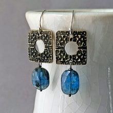 Load image into Gallery viewer, A pair of handmade dangle earrings with textured bronze square charms and blue oval gemstone beads, hanging from a white ceramic vase.