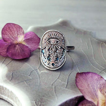 Load image into Gallery viewer, An oval silver ring featuring an intricate eye design with a botanical motif, resting on a textured gray stone surface with purple flowers as props.