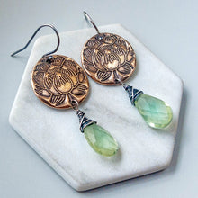 Load image into Gallery viewer, A pair of bohemian-style dangle earrings displayed on a white hexagonal ceramic tile. The earrings feature an oxidized bronze lotus disc, silver wire wrapping, and faceted light green gemstone drops.