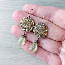 Load image into Gallery viewer, A pair of handmade dangle earrings held in a hand. The earrings feature round, antiqued bronze-toned metal discs with an engraved lotus design, hanging from silver-toned French hooks, and terminating in faceted light green gemstone drops.