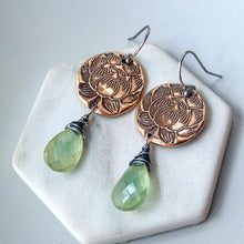 Load image into Gallery viewer, A pair of handmade dangle earrings displayed on a white hexagonal stone stand. The earrings feature round, bronze metal discs with an embossed lotus flower design and a green gemstone drop, hanging from silver-toned fishhook wires.