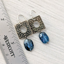 Load image into Gallery viewer, A pair of dangling earrings displayed on a white textured surface next to a ruler for scale. The earrings feature textured bronze square charms with circular cutouts and faceted blue oval gemstone beads hanging below.