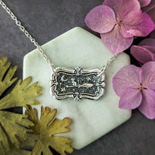 Load image into Gallery viewer, Silver necklace with decorative pendant on a white stone surface with pink flowers and green leaves.