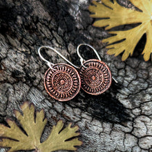 Load image into Gallery viewer, A pair of round copper-toned pendant earrings with engraved circular patterns and silver fishhook wires, displayed on textured tree bark with dried yellow leaves.