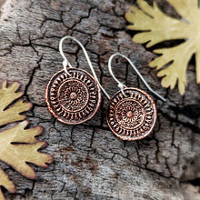 Load image into Gallery viewer, A pair of round copper-toned pendant earrings with engraved circular patterns and silver fishhook wires, displayed on textured tree bark with dried leaves.