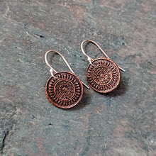 Load image into Gallery viewer, A pair of round copper-toned pendant earrings with intricate circular mandala-like embossed patterns, attached to silver fishhook wires, displayed on a dark textured slate surface.