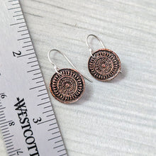 Load image into Gallery viewer, A pair of round copper-toned pendant earrings with silver fishhook wires, displayed on a light gray textured surface next to a metal ruler for scale.