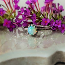 Load image into Gallery viewer, Opal ring with an intricate setting on a reflective surface with purple flowers in the background