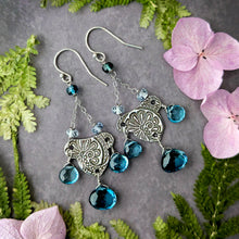 Load image into Gallery viewer, Silver earrings with London blue topaz briolettes on a gray surface with green leaves and pink flowers.