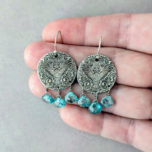 Load image into Gallery viewer, Silver earrings with an embossed owl design and blue teardrop gemstone beads held in a hand against a gray background