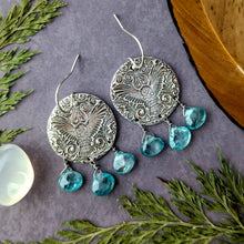 Load image into Gallery viewer, Silver earrings with an embossed owl design and blue teardrop gemstone beads on a gray surface with green leaves, a feather, and a moonstone outside of frame.