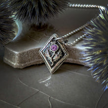 Load image into Gallery viewer, Fine silver thistle pendant with a pink sapphire on a ceramic surface with globe thistles in the foreground and background