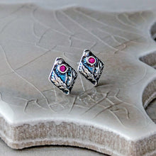 Load image into Gallery viewer, A pair of silver-toned diamond-shaped stud earrings with floral design and a small pink gemstone, displayed on a textured ceramic tile.