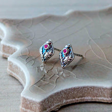 Load image into Gallery viewer, A pair of silver-toned diamond-shaped stud earrings with floral design and a small pink gemstone, displayed on a textured ceramic tile.