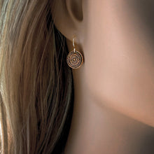 Load image into Gallery viewer, A close-up of a copper-toned circular drop earring with an intricate sunburst or filigree pattern, shown on a mannequin's ear with long light brown hair.