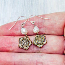 Load image into Gallery viewer, A pair of dangling earrings displayed on a person's hand. Each earring has a silver fishhook wire, a small irregular white pearl bead, and an oxidized bronze flower-shaped charm with a textured center.
