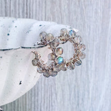Load image into Gallery viewer, A handmade wire-wrapped hoop earring featuring small grayish/beige labradorite beads with iridescent blue-green flashes, displayed on a white speckled ceramic dish against a light gray wood background.
