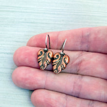 Load image into Gallery viewer, A pair of small copper drop earrings with engraved leaf-like patterns and silver hooks, resting on a person's fingers against a pale textured background.
