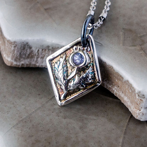 A fine silver thistle pendant with a blue tanzanite centerpiece, attached to a silver chain, displayed on a textured background.