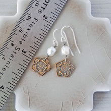 Load image into Gallery viewer, A pair of handmade dangle earrings displayed on a textured ceramic tile next to a stainless steel ruler for scale. The earrings feature silver fishhook wires, white pearl beads, and bronze flower-shaped charms.
