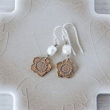 Load image into Gallery viewer, A pair of handmade dangle earrings featuring textured bronze flower charms, white pearl beads, and silver fishhook wires, displayed on a beige ceramic tile.
