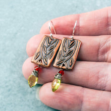 Load image into Gallery viewer, A pair of handmade dangle earrings held in a hand. The earrings feature rectangular copper plates with an embossed leaf pattern, a small orange gemstone bead, and a faceted yellow-green gemstone drop.
