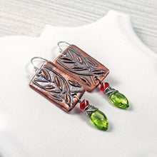 Load image into Gallery viewer, A pair of handmade dangle earrings featuring rectangular copper metal plates with engraved leaf patterns, silver fishhook wires, small red gemstone beads, and green faceted gemstones, displayed on a white surface.
