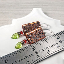 Load image into Gallery viewer, A pair of handmade rectangular drop earrings with copper metal rectangles featuring an embossed leaf pattern, silver wire hooks, small orange gemstone beads, and green faceted gemstone drops, displayed on a white ceramic dish with a stainless steel ruler for scale.
