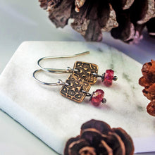 Load image into Gallery viewer, A pair of handmade dangle earrings with textured bronze square charms, silver fishhook wires, and faceted pink tourmaline beads, displayed on a white hexagonal stone tray with pine cones.
