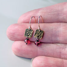 Load image into Gallery viewer, A pair of small dangling earrings held between fingers. Each earring has a square oxidized bronze-toned metal charm with an engraved pattern and a dangling faceted dark pink tourmaline bead.
