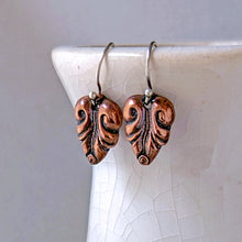 Load image into Gallery viewer, A pair of handmade dangle earrings with carved, textured copper leaf-shaped pendants on silver fishhook wires, displayed on a white ceramic surface.
