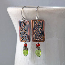 Load image into Gallery viewer, A pair of handmade rectangular drop earrings with copper metal rectangles featuring an engraved leaf pattern, silver fishhook wires, small dark orange fire opal beads, and green faceted peridot pendants, displayed on a white ceramic vase against a gray background.

