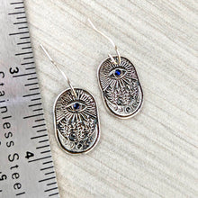 Load image into Gallery viewer, Silver eye-themed earrings on a textured surface with a ruler for scale.
