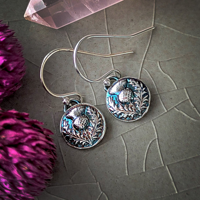 Silver earrings with intricate designs on a textured surface with purple flowers.