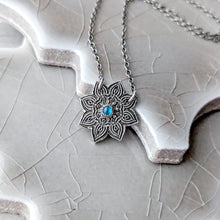 Load image into Gallery viewer, Silver mandala pendant with a blue gemstone on a chain, placed on a textured surface.
